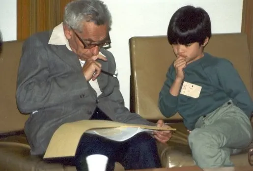 Black and white photo of Paul Erdős, an older man in glasses, sitting at a table with a young Terence Tao, about ten years old, working through a book together.
