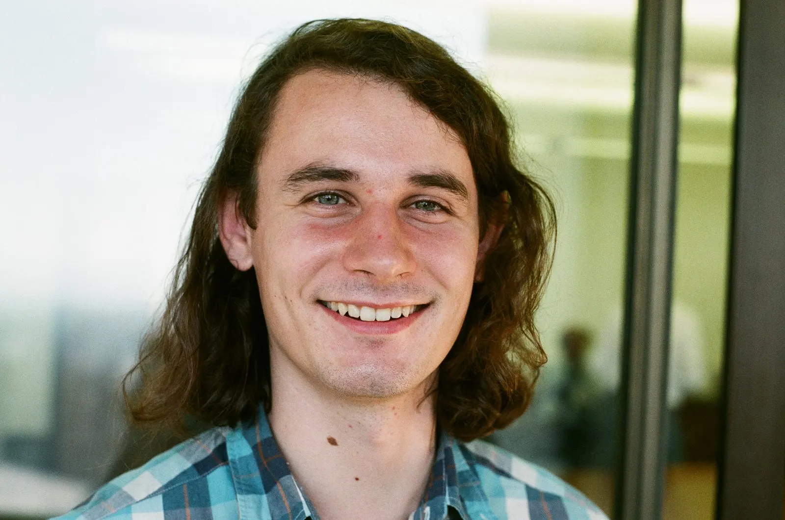 Portrait of Peter Scholze, a young man with shoulder-length brown hair and a plaid shirt, smiling
