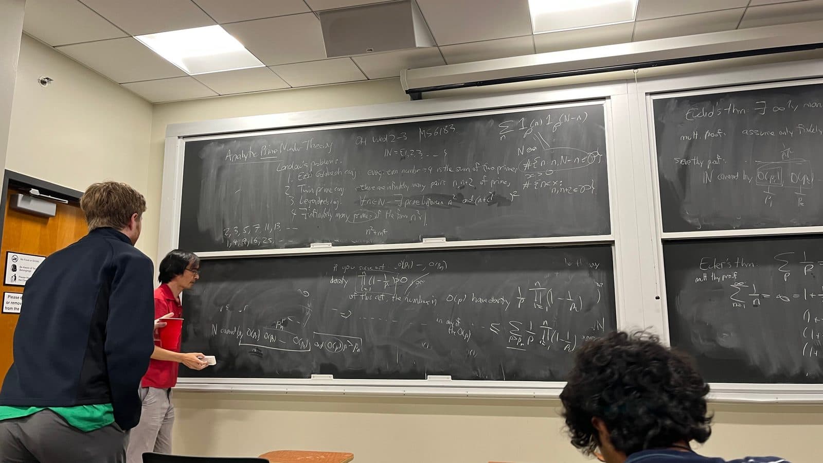Terence Tao in a red shirt writing at a chalkboard covered in analytic number theory, with students watching